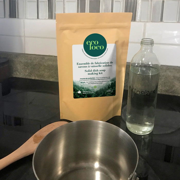 Pochette en papier kraft avec le logo d'Eco Loco contenant un ensemble de fabrication de savons à vaisselle solides sur une comptoir de cuisine avec un chaudron en métal, une bouteille en verre vitre contenant du vinaigre et une cuillère en bois