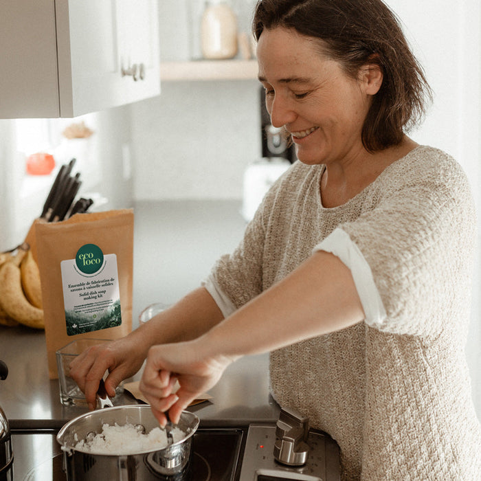 Femme qui sourit en mélangeant le contenant d'une pochette en papier kraft avec le logo d'Eco Loco contenant un ensemble de fabrication de savons à vaisselle solides dans une cuisine blanche lumineuse avec des bananes sur le comptoir
