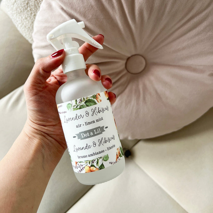 Hand holding a bottle of Dot & Lil air freshener with a floral label against a neutral background.